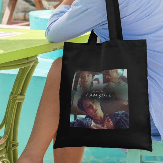 Woman sitting next to table with black totebag with i am still jungkook image design over shoulders