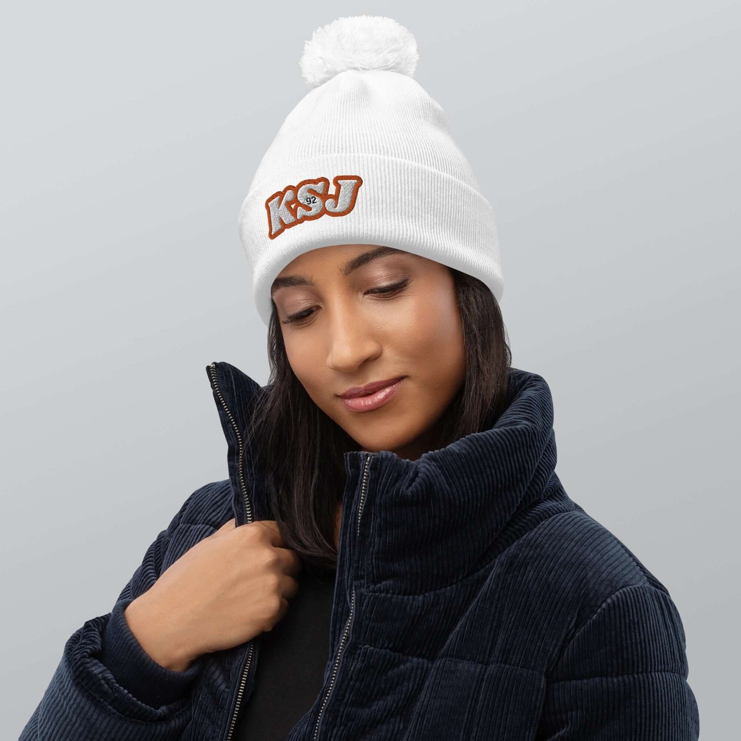 Woman wearing a white beanie with pom pom and KSU logo, cozy in a corduroy jacket.