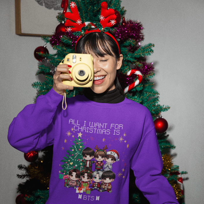 Woman wearing BTS Chibi Holiday Magic Sweater, smiling with a camera, in front of a Christmas tree with antler headband.