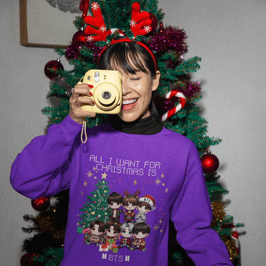 Woman wearing BTS Chibi Holiday Magic Sweater, smiling with a camera, in front of a Christmas tree with antler headband.